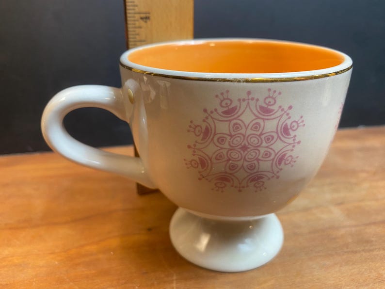 K&ouml;nnte beinhalten: Cremefarbene Teetasse mit wei&szlig;em Henkel und Sockel. Der Innenraum ist orange und der Rand ist goldfarben verziert. Ein rosa Blumendesign verziert die Au&szlig;enseite. Eine dekorative Teetasse zum Servieren von Tee oder Kaffee.