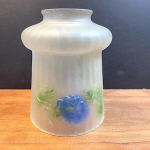 Może przedstawiać: Mrożony szklany klosz lampy z rozkloszowaną górą i cylindrycznym korpusem. Klosz ma niebieski kwiatowy wzór z zielonymi liśćmi. Klosz lampy ma rozpraszać światło i dodawać dekoracyjny akcent.