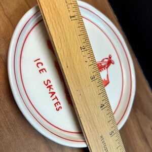 May include: A small, round, white plate with a red rim and the words "ICE SKATES" in red lettering. A wooden ruler, marked in inches, is placed across the plate. The plate features a red graphic of ice skates.