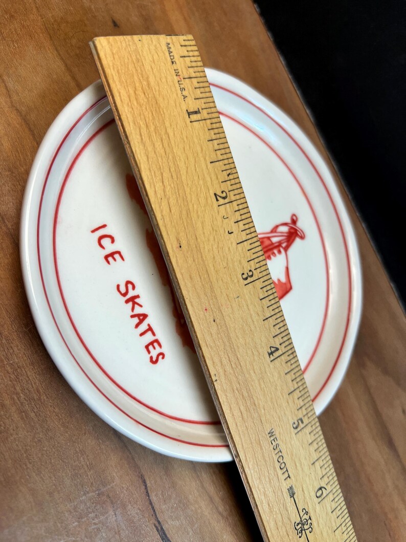 May include: A small, round, white plate with a red rim and the words "ICE SKATES" in red lettering. A wooden ruler, marked in inches, is placed across the plate. The plate features a red graphic of ice skates.
