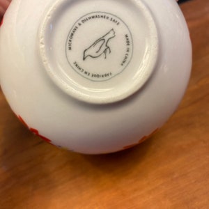 K&ouml;nnte beinhalten: Wei&szlig;er Keramiknapf mit farbenfrohem Blumendesign. Der Boden der Sch&uuml;ssel zeigt eine schwarze Vogelillustration in einem Kreis mit dem Text "MICROWAVE & DISHWASHER SAFE" und "MADE IN CHINA".