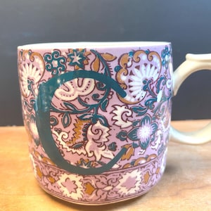 May include: A ceramic mug featuring a floral design in pink, white, teal, and gold. A teal "C" is displayed on the mug. The handle is off-white.