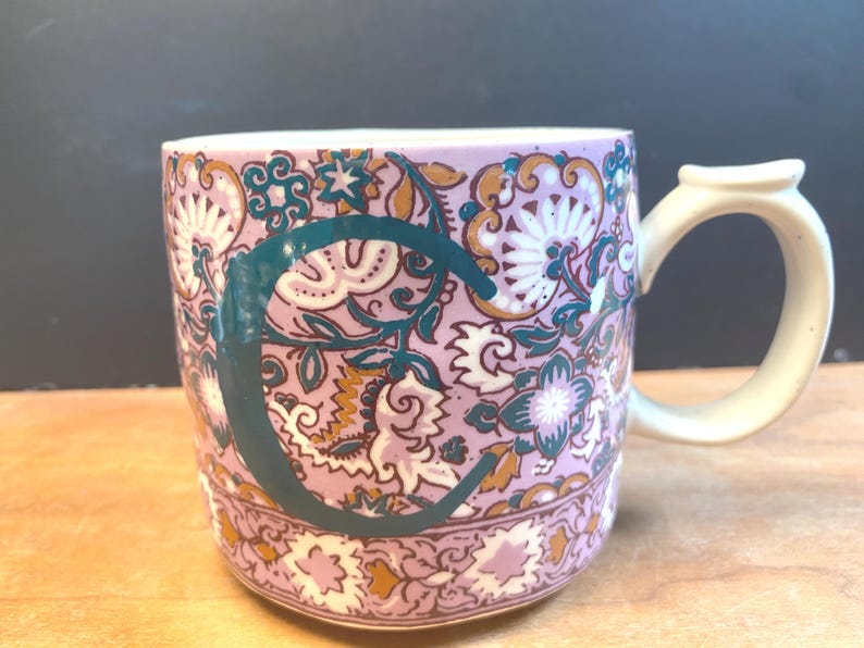 May include: A ceramic mug featuring a floral design in pink, white, teal, and gold. A teal "C" is displayed on the mug. The handle is off-white.