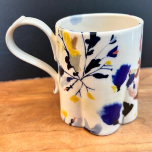 May include: A white ceramic mug with a unique, scalloped base and a curved handle. The mug is decorated with a floral pattern in shades of blue, yellow, and brown. The mug is sitting on a wooden surface.