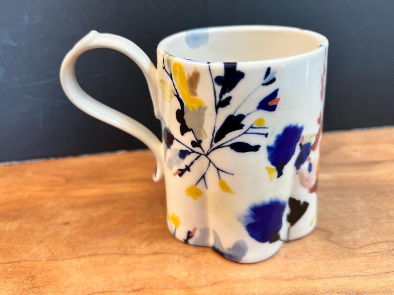 May include: A white ceramic mug with a unique, scalloped base and a curved handle. The mug is decorated with a floral pattern in shades of blue, yellow, and brown. The mug is sitting on a wooden surface.