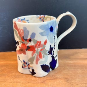 May include: A white ceramic mug with a floral design in shades of red, blue, and gray. The mug has a curved handle and a slightly irregular shape. The floral pattern covers the exterior of the mug.