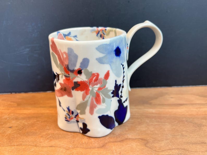 May include: A white ceramic mug with a floral design in shades of red, blue, and gray. The mug has a curved handle and a slightly irregular shape. The floral pattern covers the exterior of the mug.