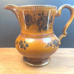 May include: A vintage ceramic jug with a warm, amber glaze. The jug features black floral designs and a curved handle. The rim and base have a subtle, metallic trim. This decorative jug is a classic piece for serving beverages.