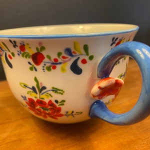 K&ouml;nnte beinhalten: Wei&szlig;e Keramik-Teetasse mit blauem Henkel und Rand, verziert mit einem Blumenmuster in Rot, Gelb und Blau. Der Henkel hat einen rot-wei&szlig;en Akzent, der dem Design einen Hauch von Laune verleiht.