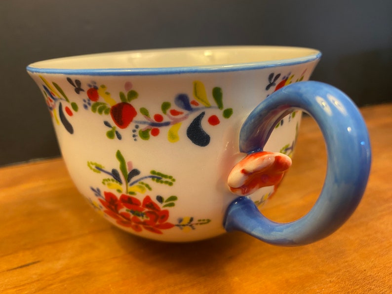 K&ouml;nnte beinhalten: Wei&szlig;e Keramik-Teetasse mit blauem Henkel und Rand, verziert mit einem Blumenmuster in Rot, Gelb und Blau. Der Henkel hat einen rot-wei&szlig;en Akzent, der dem Design einen Hauch von Laune verleiht.