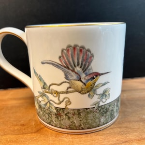 Może przedstawiać: Biały ceramiczny kubek z kolorową ilustracją kolibra siedzącego na winorośli. Kubek posiada złocony brzeg i uchwyt oraz ozdobny zielono-szary marmurkowy pasek wokół podstawy, co stanowi wyraźny i szczegółowy akcent wizualny całego naczynia.