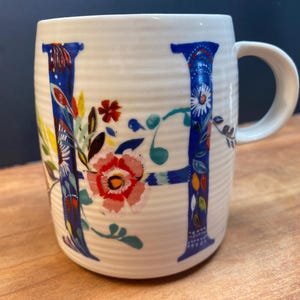 Può includere: Tazza in ceramica bianca con un design floreale blu a forma di "H". La lettera è adornata da fiori e foglie colorate. La tazza ha un manico curvo e una texture a coste. Il design include fiori rossi, rosa e gialli.