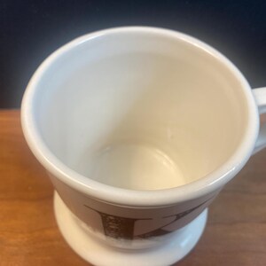 May include: Cream-colored ceramic mug with a handle and a pedestal base. The mug features a brown letter "K" design on the lower portion. The mug is empty and sits on a wooden surface.