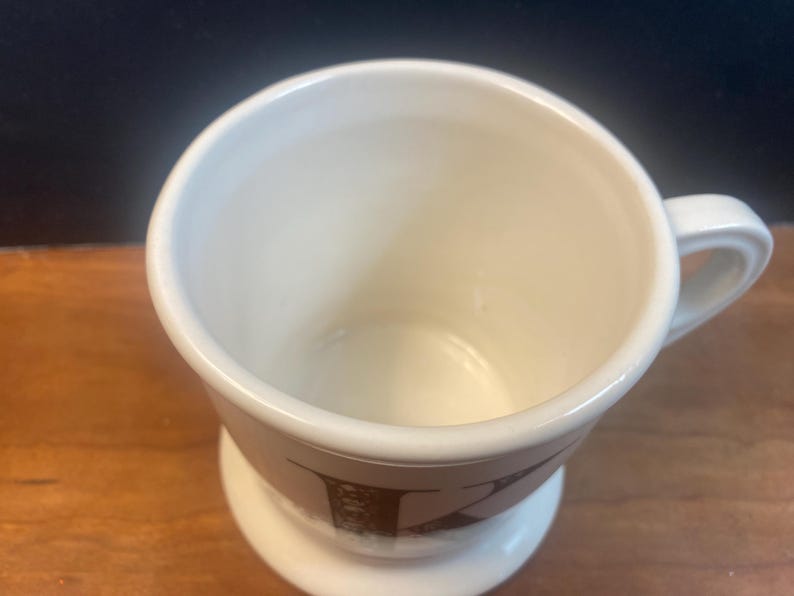 May include: Cream-colored ceramic mug with a handle and a pedestal base. The mug features a brown letter "K" design on the lower portion. The mug is empty and sits on a wooden surface.
