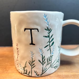 Può includere: Una tazza di ceramica bianca con manico nero e la scritta "But First, Coffee" in grassetto nero. La tazza è posta su uno sfondo neutro, che ne evidenzia il design semplice. La tazza è alta circa 10 cm.