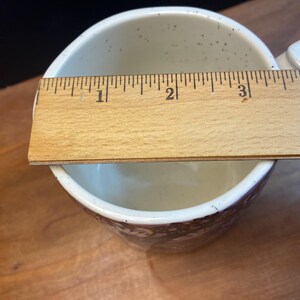 May include: Cream ceramic mug with a brown floral pattern around the base. The mug has a handle and is approximately 10 cm in diameter. The interior has small brown speckles. A wooden ruler is placed across the top of the mug.