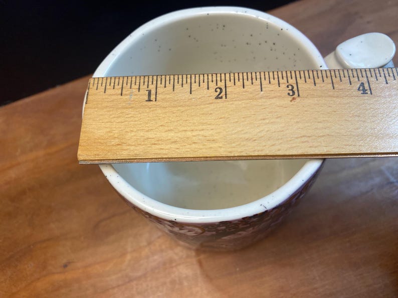 May include: Cream ceramic mug with a brown floral pattern around the base. The mug has a handle and is approximately 10 cm in diameter. The interior has small brown speckles. A wooden ruler is placed across the top of the mug.