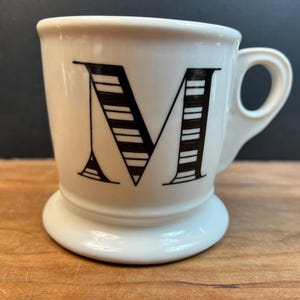 Puede incluir: Una taza de cer&aacute;mica blanca con la letra "M" negra en la parte delantera. La letra est&aacute; estilizada con rayas horizontales. La taza tiene un asa peque&ntilde;a y una base redondeada, colocada sobre una superficie de madera.
