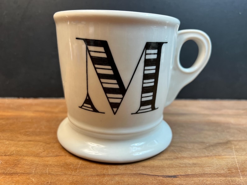 Puede incluir: Una taza de cer&aacute;mica blanca con la letra "M" negra en la parte delantera. La letra est&aacute; estilizada con rayas horizontales. La taza tiene un asa peque&ntilde;a y una base redondeada, colocada sobre una superficie de madera.