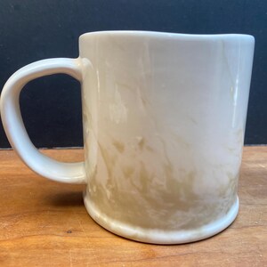 Puede incluir: Taza de cer&aacute;mica color crema con asa redondeada. La taza tiene un efecto marmolado con tonos marr&oacute;n claro y beige. La taza est&aacute; sobre una superficie de madera con un fondo oscuro.