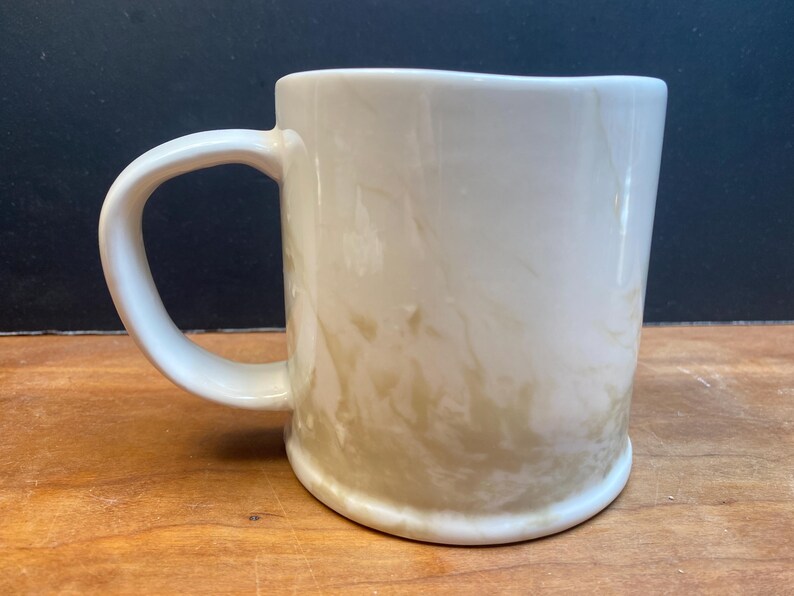 Puede incluir: Taza de cer&aacute;mica color crema con asa redondeada. La taza tiene un efecto marmolado con tonos marr&oacute;n claro y beige. La taza est&aacute; sobre una superficie de madera con un fondo oscuro.