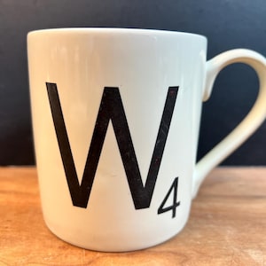 Puede incluir: Una taza de cerámica blanca con la letra negra "W" y el número "4" impresos en la parte delantera. La taza tiene un asa curva y está sobre una superficie de madera.