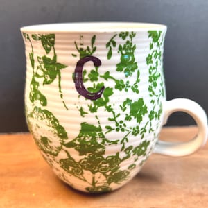 May include: White ceramic mug with a green floral pattern and a purple letter "C". The mug has a rounded shape with a white handle. The floral design covers the exterior of the mug.