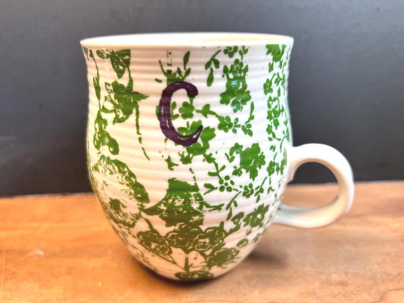 May include: White ceramic mug with a green floral pattern and a purple letter "C". The mug has a rounded shape with a white handle. The floral design covers the exterior of the mug.
