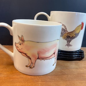 May include: Two white ceramic mugs with illustrations. One mug features a pink pig, the other a rooster with a red comb.