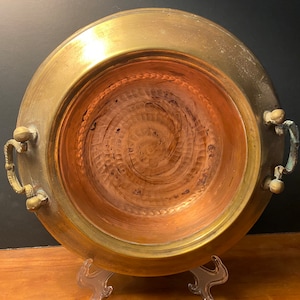 May include: A large, round, brass serving bowl with two handles. The bowl has a copper interior with a hammered, swirling pattern. The bowl is on a white stand.