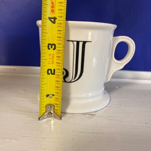 Może przedstawiać: Biały ceramiczny kubek z czarną literą "J" z przodu. Kubek ma około 10 cm wysokości. Ma uchwyt i lekko rozszerzoną krawędź. Kubek stoi na białej powierzchni.