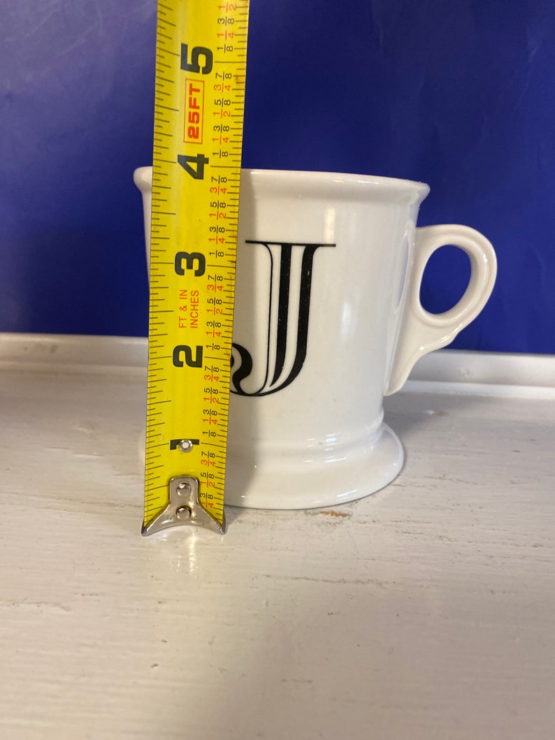 Może przedstawiać: Biały ceramiczny kubek z czarną literą "J" z przodu. Kubek ma około 10 cm wysokości. Ma uchwyt i lekko rozszerzoną krawędź. Kubek stoi na białej powierzchni.