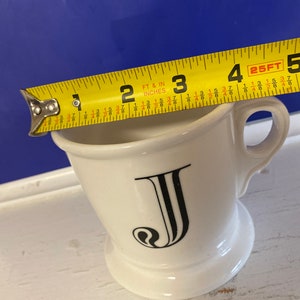 Może przedstawiać: Biały ceramiczny kubek do golenia z czarną literą "J" z przodu. Kubek ma około 10 cm wysokości i ma uchwyt. Ż&oacute;łta miarka jest umieszczona na g&oacute;rze kubka.