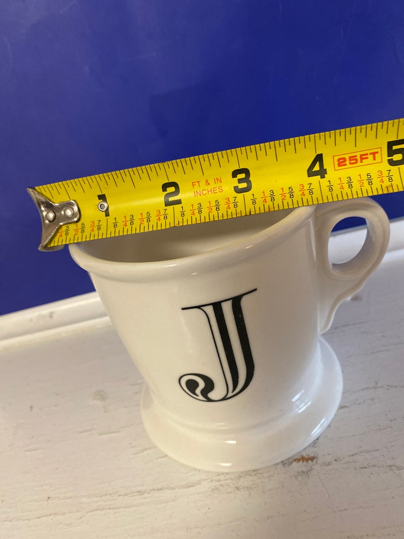 Może przedstawiać: Biały ceramiczny kubek do golenia z czarną literą "J" z przodu. Kubek ma około 10 cm wysokości i ma uchwyt. Ż&oacute;łta miarka jest umieszczona na g&oacute;rze kubka.