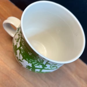 May include: A white ceramic mug with a green tree-like pattern wraps around the exterior. The mug has a curved handle and a cream-colored interior. The mug is sitting on a wooden surface.
