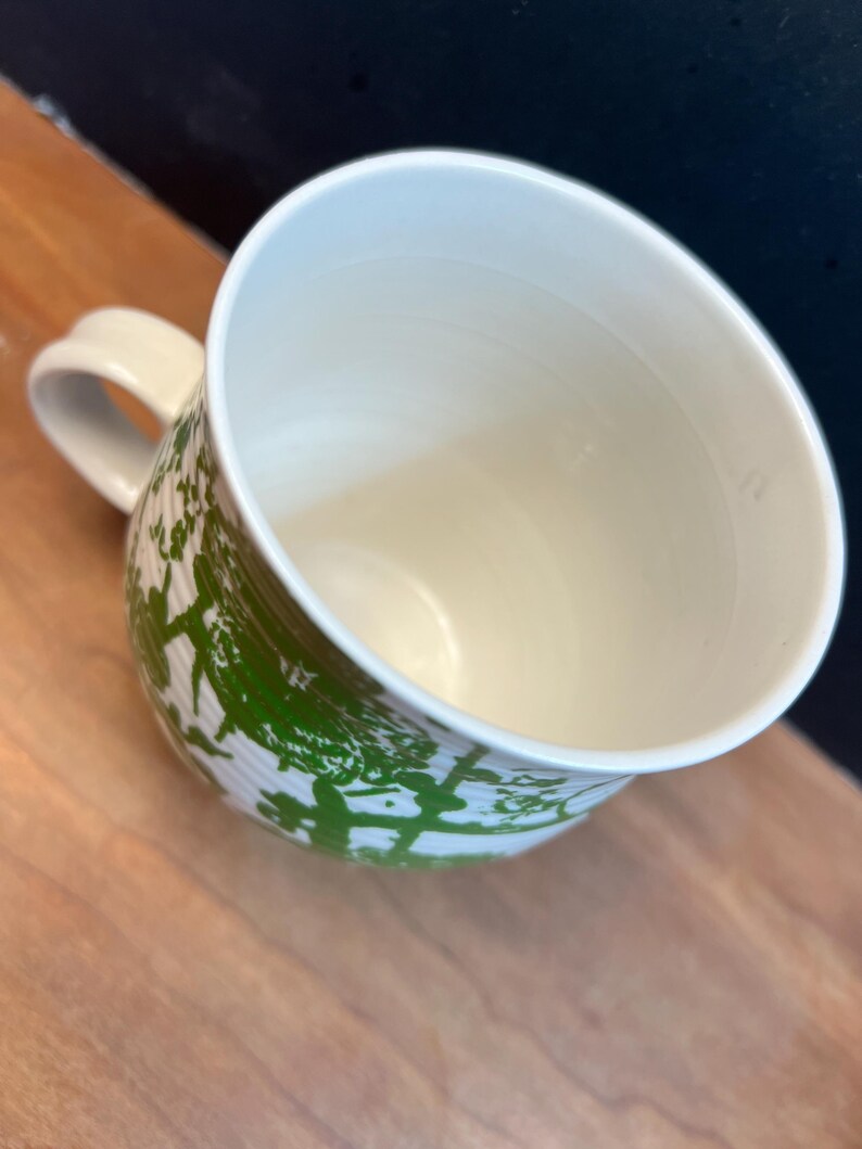 May include: A white ceramic mug with a green tree-like pattern wraps around the exterior. The mug has a curved handle and a cream-colored interior. The mug is sitting on a wooden surface.