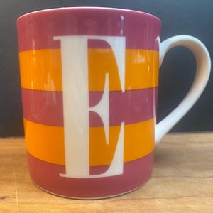 May include: A ceramic mug with a white handle features a large white letter "E" centered on a design of horizontal stripes. The stripes alternate between shades of orange and a deep pink color.