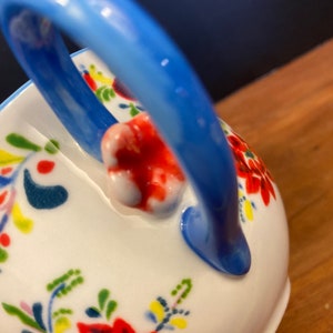 K&ouml;nnte beinhalten: Wei&szlig;e Keramikservierschale mit blauem Griff und floralem Design. Die Schale hat ein lebendiges Muster aus roten, gr&uuml;nen, gelben und blauen Blumen und Bl&auml;ttern. Der Griff ist ein leuchtendes, einfarbiges Blau.