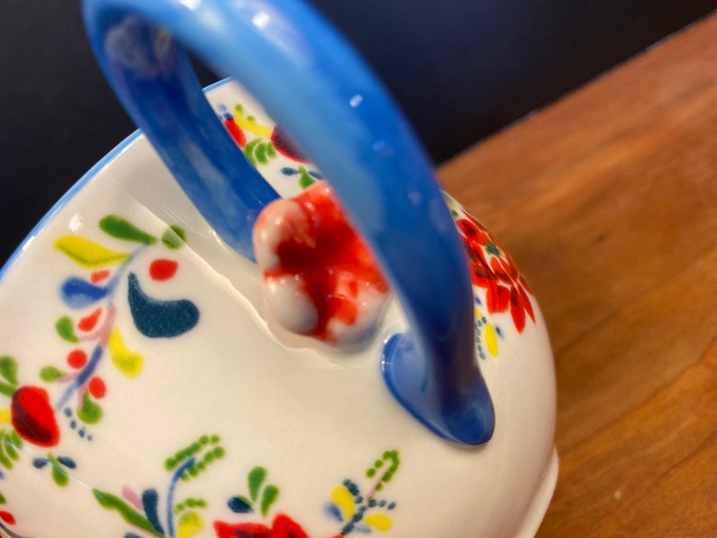 K&ouml;nnte beinhalten: Wei&szlig;e Keramikservierschale mit blauem Griff und floralem Design. Die Schale hat ein lebendiges Muster aus roten, gr&uuml;nen, gelben und blauen Blumen und Bl&auml;ttern. Der Griff ist ein leuchtendes, einfarbiges Blau.