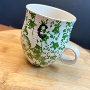 May include: White ceramic mug with a green floral pattern and a purple letter "C". The mug has a curved shape and a white handle. The mug is sitting on a wooden surface.