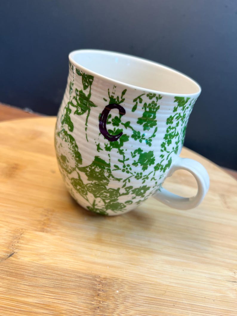 May include: White ceramic mug with a green floral pattern and a purple letter "C". The mug has a curved shape and a white handle. The mug is sitting on a wooden surface.