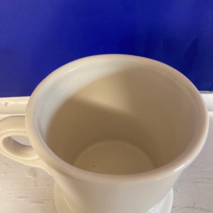 Może przedstawiać: Biały ceramiczny kubek z uchwytem. Kubek ma szeroki otw&oacute;r i zaokrągloną podstawę. Wnętrze kubka jest widoczne. Tło jest jednolicie niebieskie.