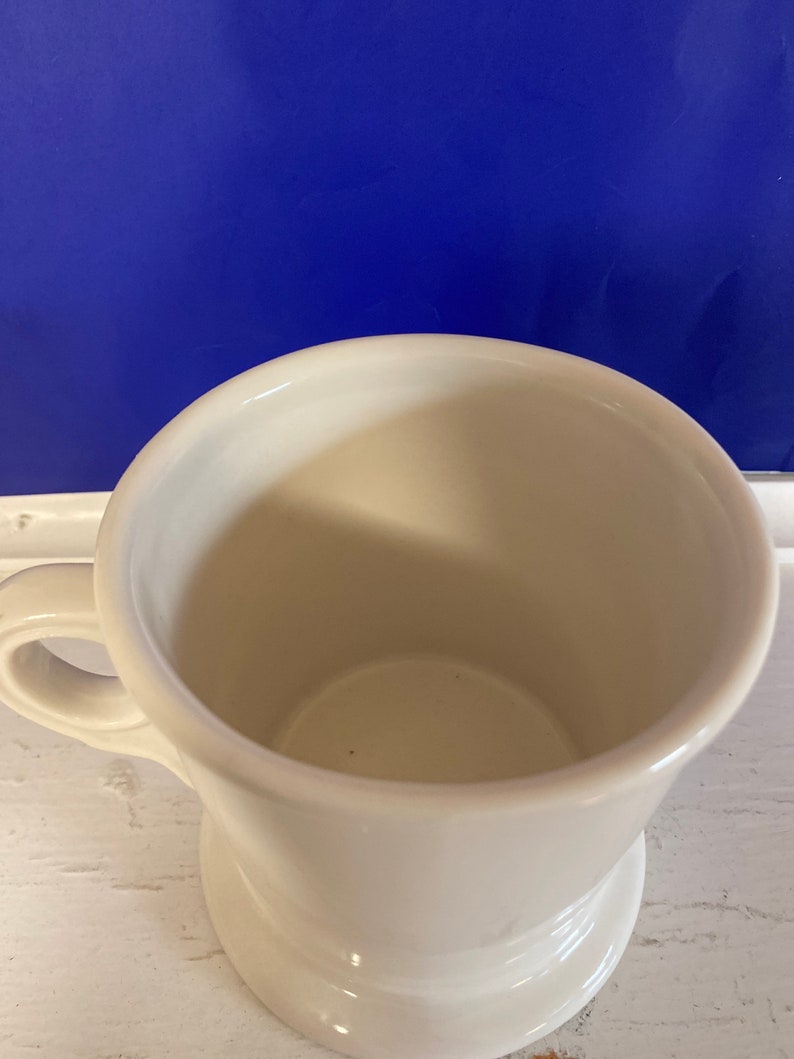 Może przedstawiać: Biały ceramiczny kubek z uchwytem. Kubek ma szeroki otw&oacute;r i zaokrągloną podstawę. Wnętrze kubka jest widoczne. Tło jest jednolicie niebieskie.