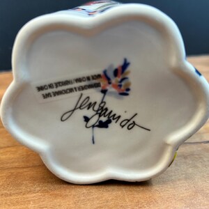 May include: White ceramic floral-shaped object with colorful accents. The bottom features a signature and text that reads "DISHWASHER & MICROWAVE SAFE MADE IN CHINA/FABRIQUE EN CHINE".