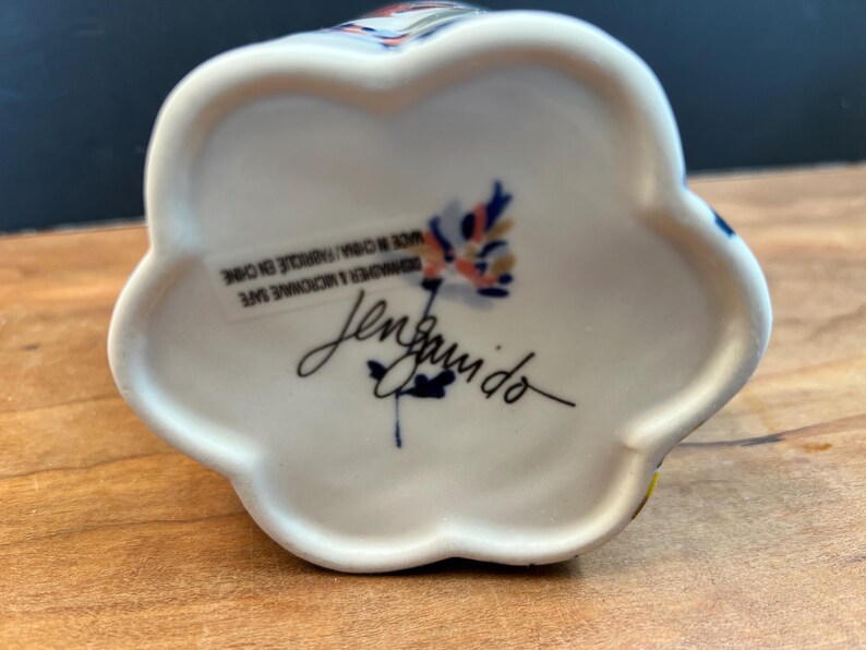 May include: White ceramic floral-shaped object with colorful accents. The bottom features a signature and text that reads "DISHWASHER & MICROWAVE SAFE MADE IN CHINA/FABRIQUE EN CHINE".