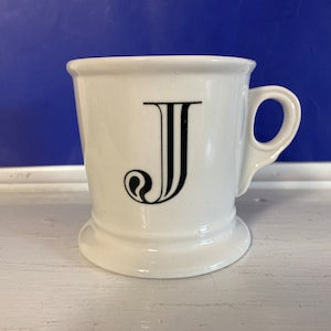 Może przedstawiać: Biały ceramiczny kubek z czarną literą "J" z przodu. Kubek ma uchwyt i rozszerzaną podstawę. Tło jest w jednolitym niebieskim kolorze. Kubek stoi na białej powierzchni.