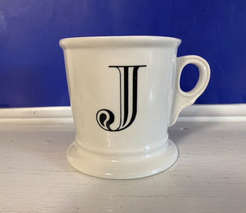 Może przedstawiać: Biały ceramiczny kubek z czarną literą "J" z przodu. Kubek ma uchwyt i rozszerzaną podstawę. Tło jest w jednolitym niebieskim kolorze. Kubek stoi na białej powierzchni.
