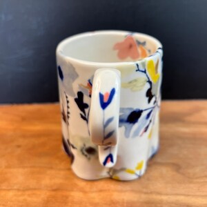 May include: A white ceramic mug with a floral design. The mug features a handle and is decorated with blue, yellow, pink, and black floral patterns. The mug is sitting on a wooden surface.