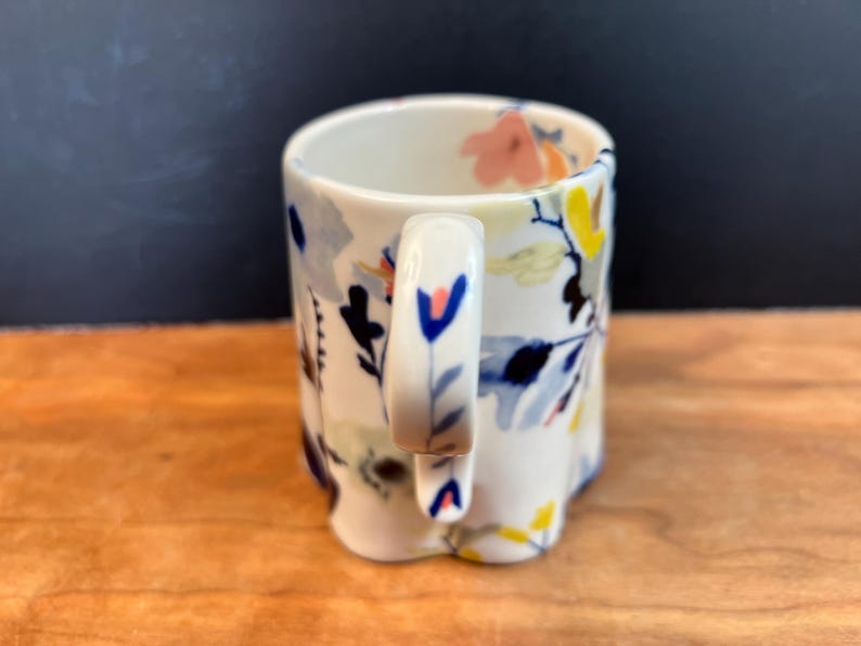 May include: A white ceramic mug with a floral design. The mug features a handle and is decorated with blue, yellow, pink, and black floral patterns. The mug is sitting on a wooden surface.