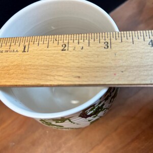 May include: A white ceramic bowl with a decorative green and brown pattern around the base. A wooden ruler, marked in inches, is placed across the top of the bowl, measuring approximately 3 inches in diameter.